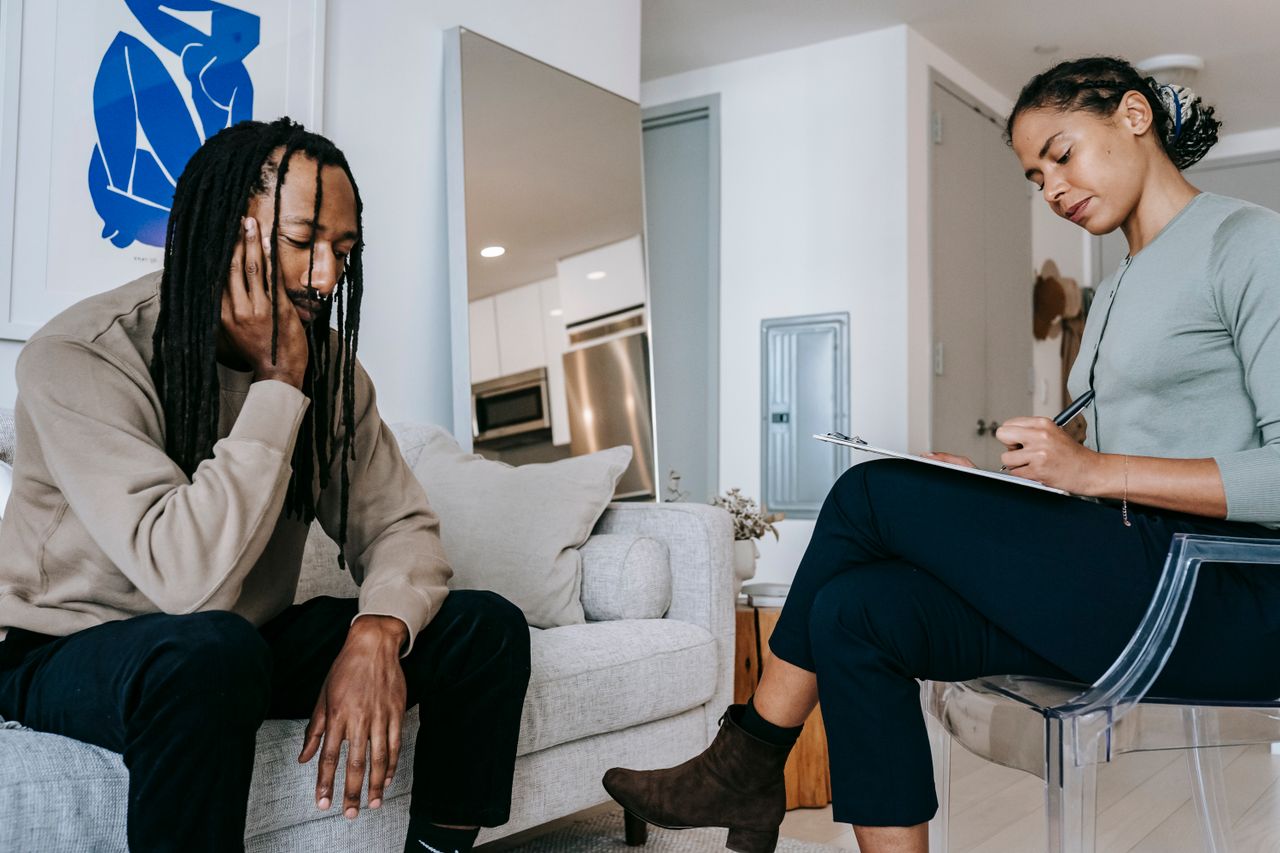 Home Attentive African American female with clipboard taking notes near sad male partner touching face on couch at home